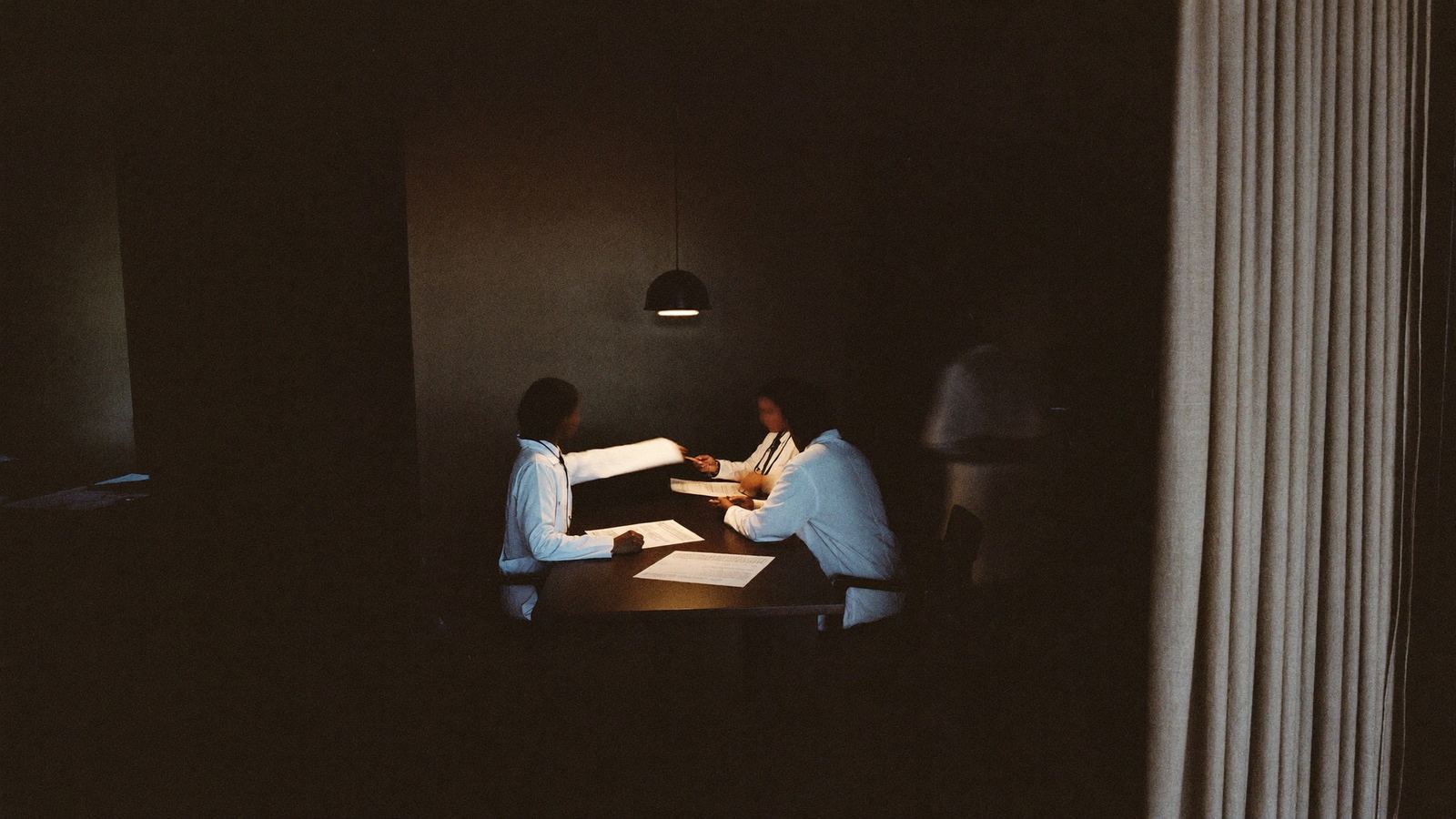 Dos personas de bata revisando papeles en una mesa, bajo una lámpara